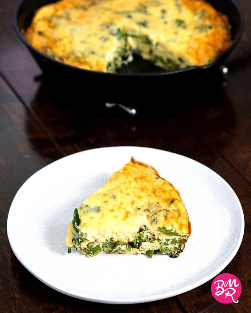slice of broccoli cheese frittata on a white plate in front of the rest of the frittata blurred in the background in a cast iron pan