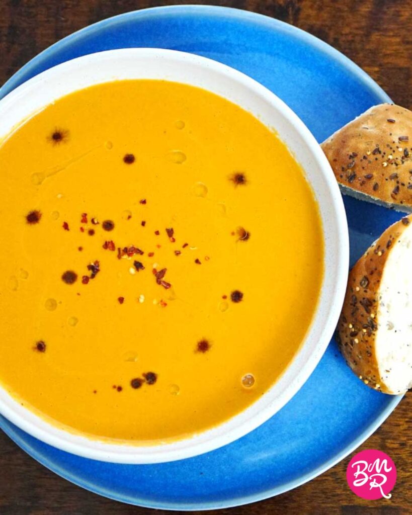 overhead view of a bowl of roasted tomato soup placed on a blue plate served with two slices of crusty seeded bread on a dark wooden surface
